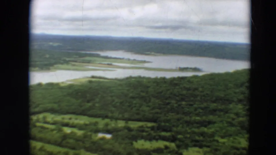 Aerial 8mm view from 1967 Minnesota showing rolling green woodland, a broad waterway, and scattered light clearings.