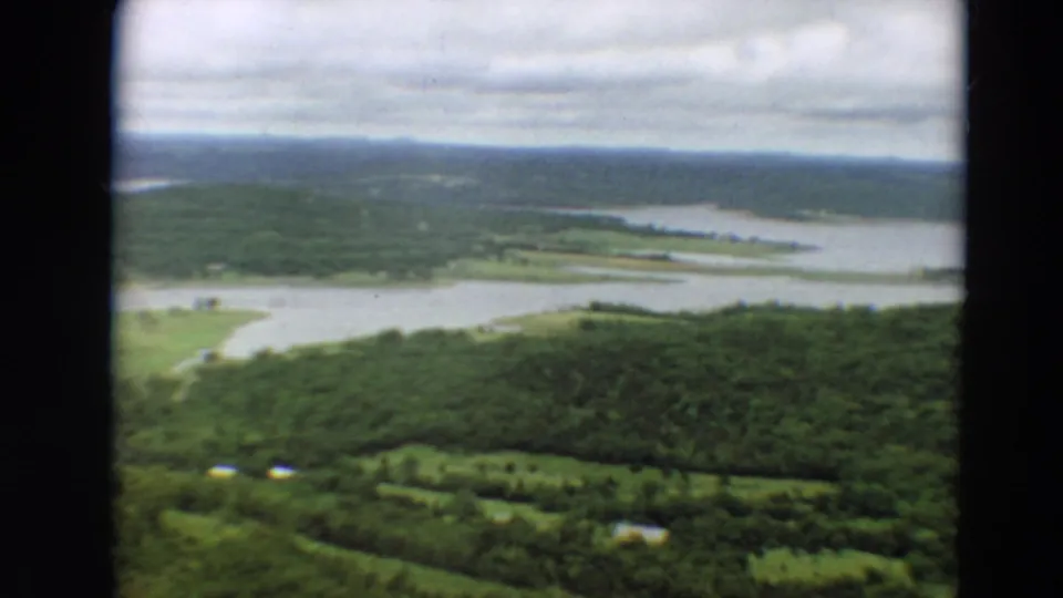 1967 Minnesota aerial frame showing forested land, open water, pale fields or marshland, and a cloudy horizon.