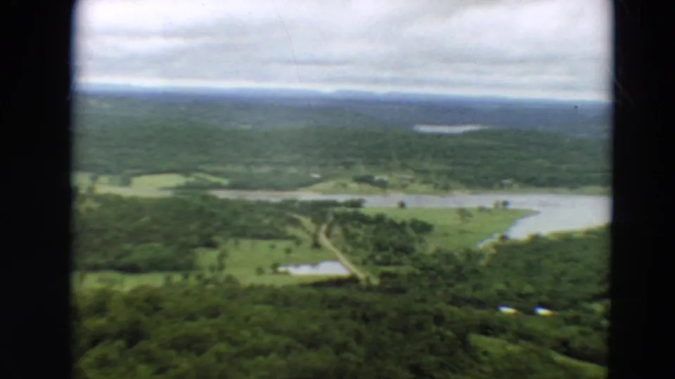 1967 Minnesota 8mm frame looking across rural green land with broad water, field patches, wooded areas, and a cloudy horizon.