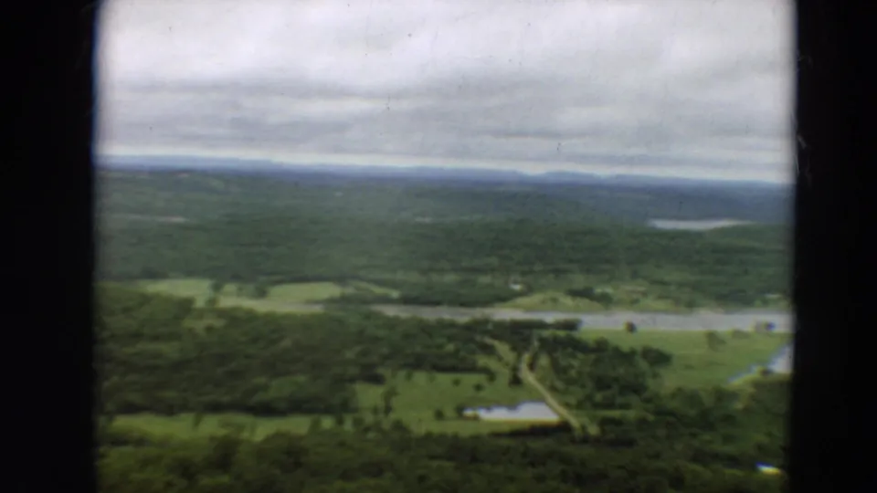 1967 Minnesota aerial frame showing fields, wooded land, water, and small distant buildings within a broad rural landscape.