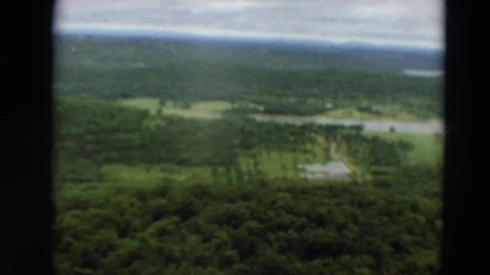 1967 Minnesota 8mm aerial view over forested land with distant water, grassy openings, and dark aircraft-window framing.