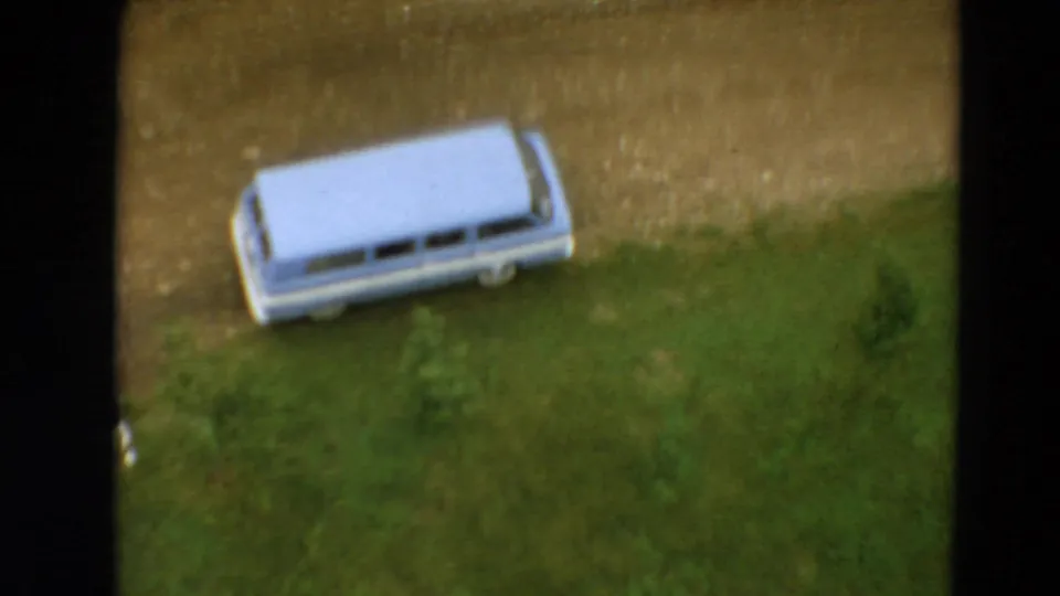 Elevated 1967 Minnesota 8mm view of the parked light blue station wagon, grass, shrubs, and a small person near the lower frame edge.