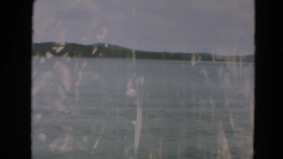 1967 Minnesota 8mm frame showing calm lake water and distant green hills beneath an overcast sky, with visible film wear.