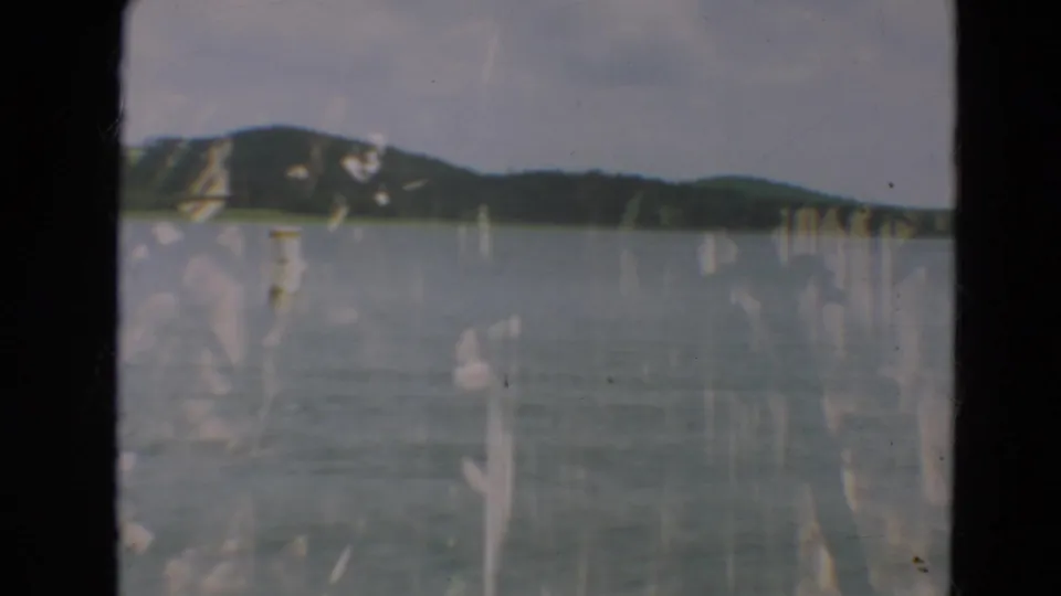 1967 Minnesota lake scene with low wooded hills across the water, muted sky, and prominent 8mm film wear marks.