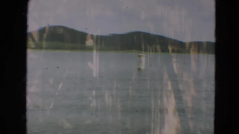 Minnesota 1967 home-movie frame of a calm lake below rolling green hills, softened by grain and vertical film streaking.