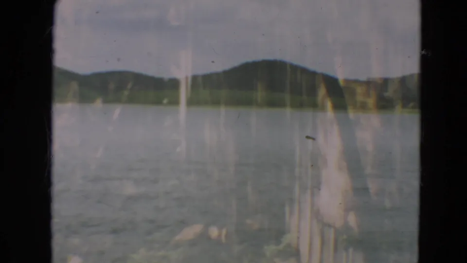 1967 Minnesota 8mm view across still lake water toward wooded hills, with soft exposure and visible physical-film texture.