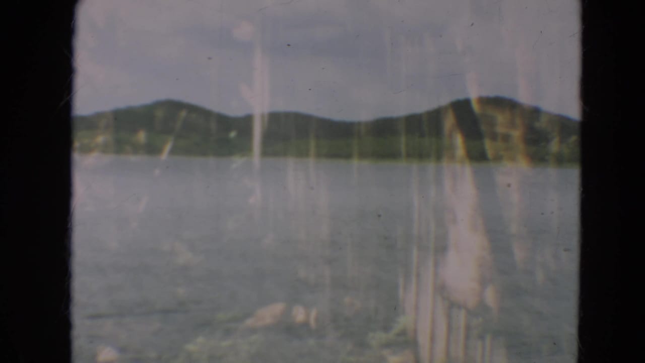 1967 Minnesota 8mm frame showing a calm lake, low wooded hills, pale sky, and dark vertical frame edges.