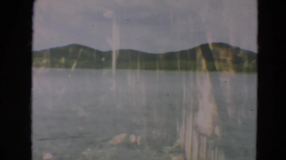 1967 Minnesota 8mm frame showing a calm lake, low wooded hills, pale sky, and dark vertical frame edges.