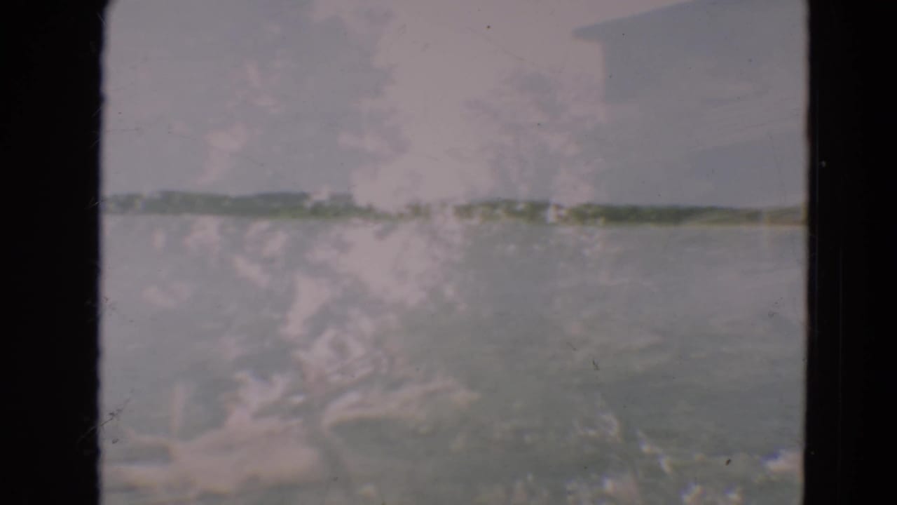 1967 8mm frame from Minnesota showing a soft, faded lakeside view with water, distant shore, and heavy film gate borders.