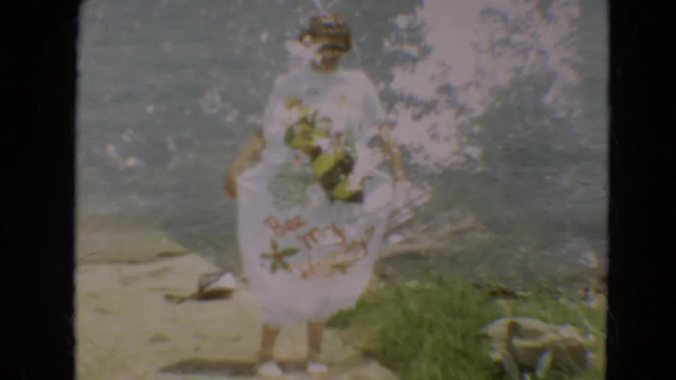 1967 Minnesota home-movie frame showing a woman posing by the lake in sunglasses and a white dress with bee-themed text.