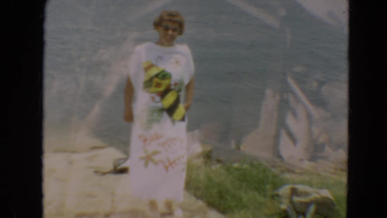 Adult woman in sunglasses stands on a Minnesota shoreline in a white beach cover-up during 1967 8mm home-movie footage.