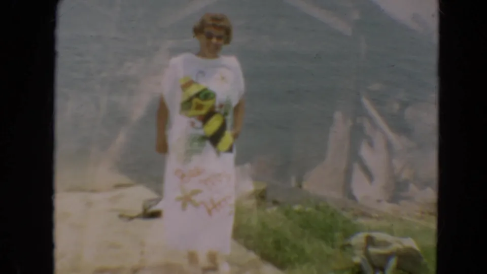 Adult woman in sunglasses stands on a Minnesota shoreline in a white beach cover-up during 1967 8mm home-movie footage.