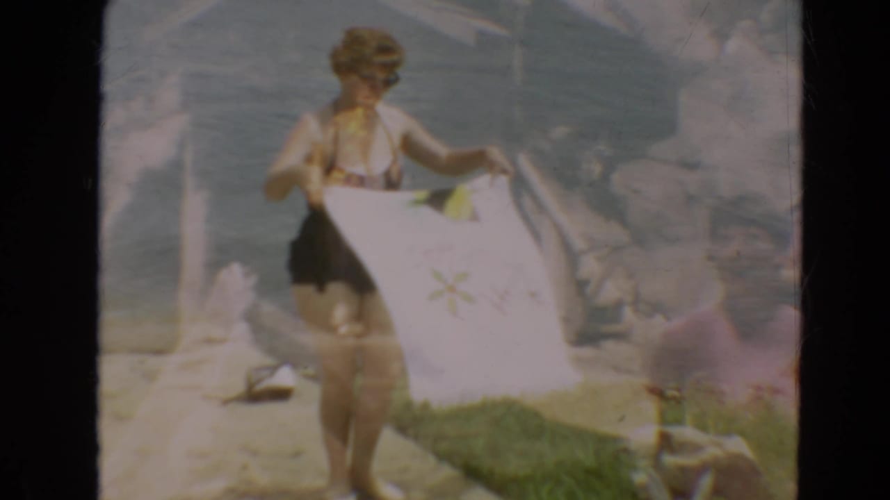 Woman holding a white cover-up open while standing by the water in a dark swimsuit.