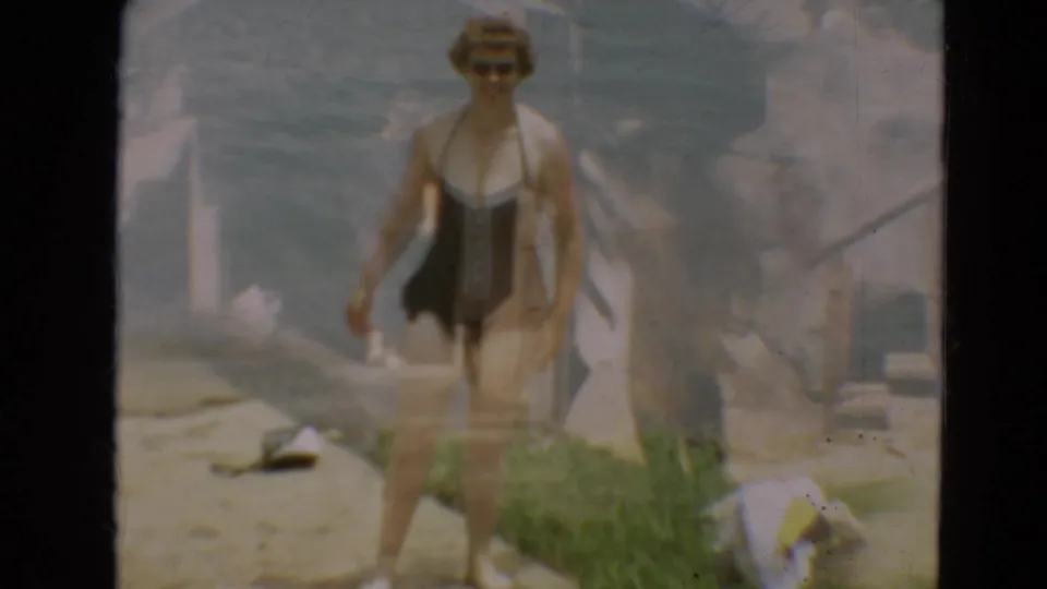 Adult woman wearing sunglasses and a dark one-piece swimsuit walks toward the camera beside water in 1967 Minnesota home-movie footage.