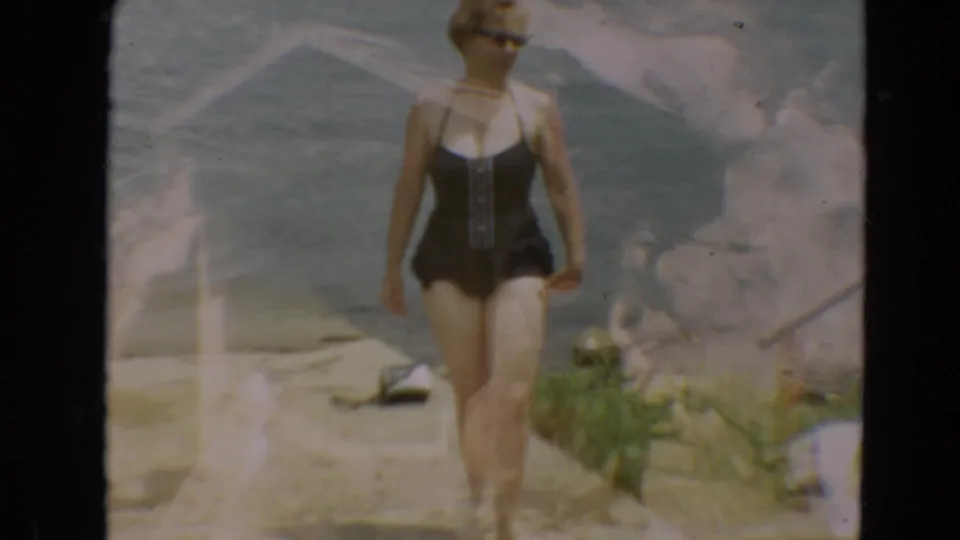 Adult woman in sunglasses and a navy one-piece swimsuit stands on a lakeshore in 1967 Minnesota 8mm home-movie footage.