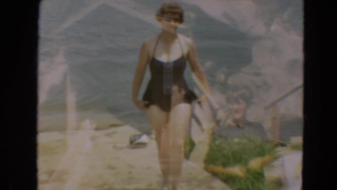 Woman in black swimsuit and sunglasses walking beside a lake.