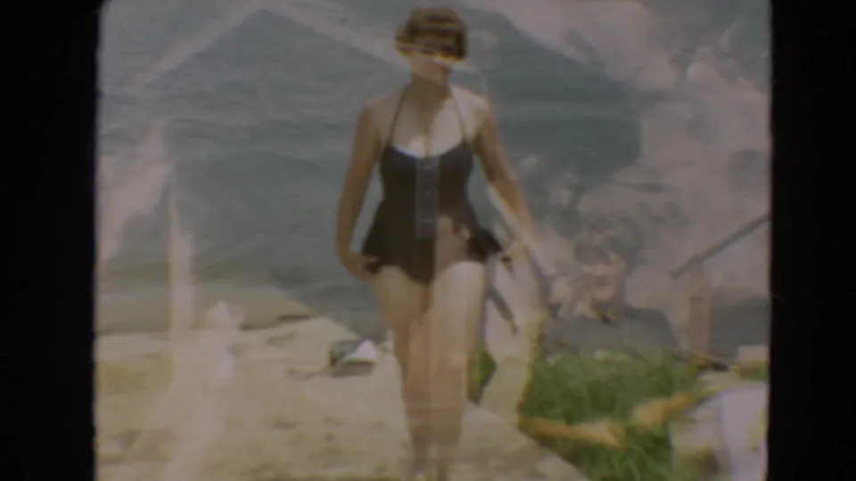 In 1967 Minnesota home-movie footage, a woman in a black one-piece swimsuit walks along a lakeside concrete edge.