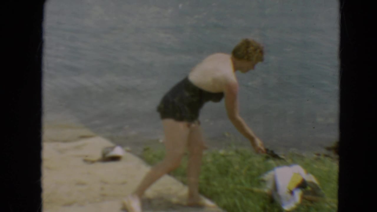 The woman bends near personal items on the grass and concrete beside the Minnesota lakeshore.