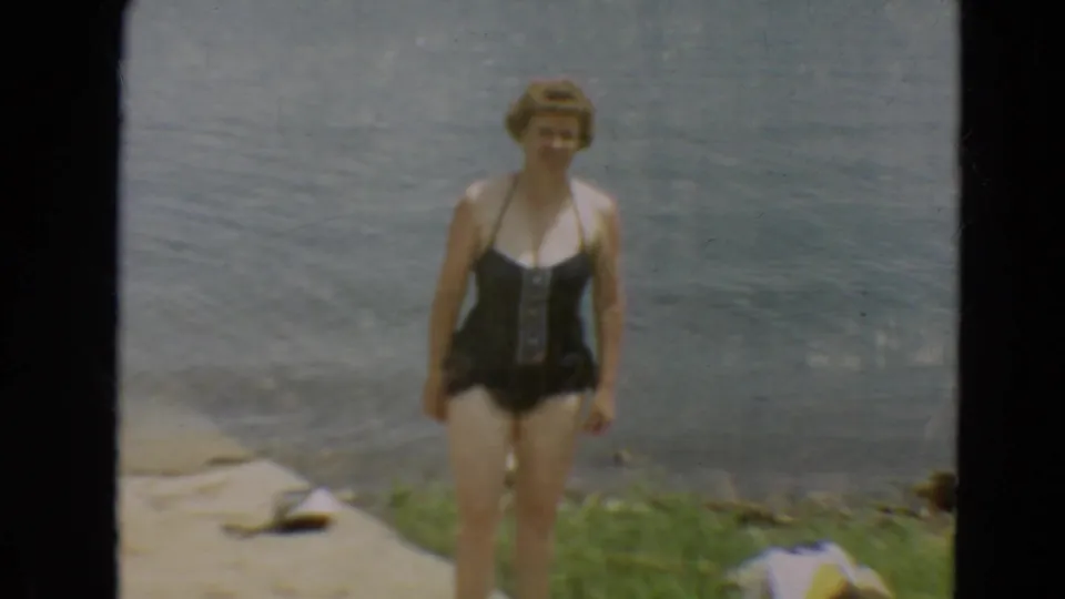 A woman in a black one-piece swimsuit stands facing the camera with lake water behind her in 1967 Minnesota footage.