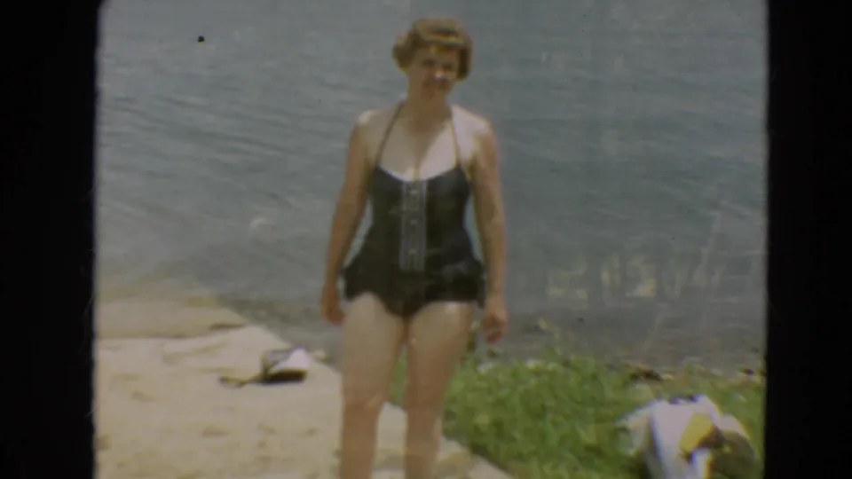 The woman walks toward the camera along the lakeside edge, with water, grass, and personal items visible nearby.