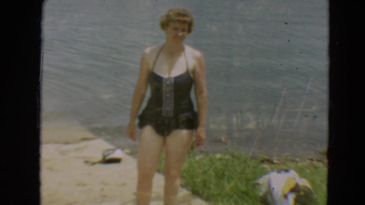 1967 Minnesota home-movie frame showing a woman in a dark one-piece swimsuit facing the camera beside calm lake water.