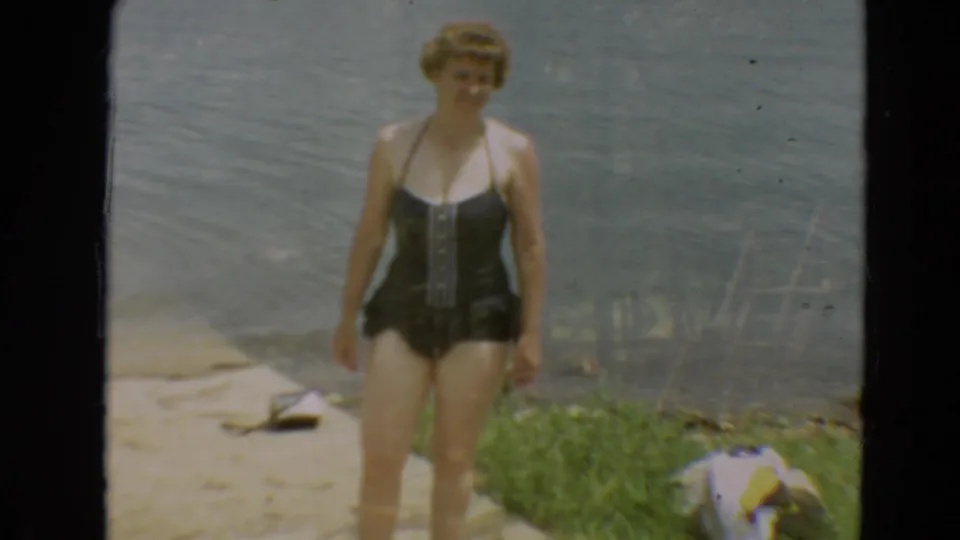 1967 Minnesota home-movie frame showing a woman in a dark one-piece swimsuit facing the camera beside calm lake water.