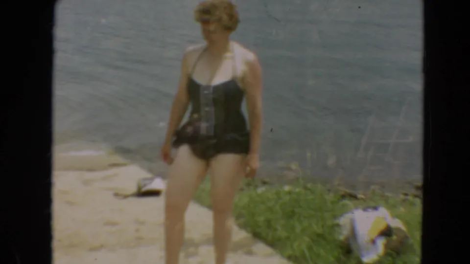 1967 Minnesota lakeside frame with a woman in a dark swimsuit walking near the water and grassy bank.