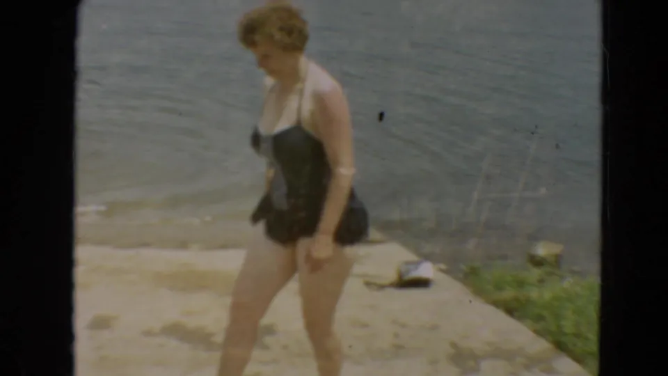 1967 Minnesota home-movie still of a woman in a dark swimsuit passing along a lakeside path with water behind her.