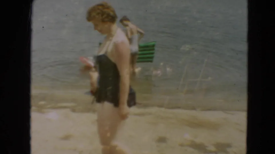 1967 Minnesota lakeside frame showing a woman in a dark swimsuit in the foreground and another swimmer with a green towel in shallow water.