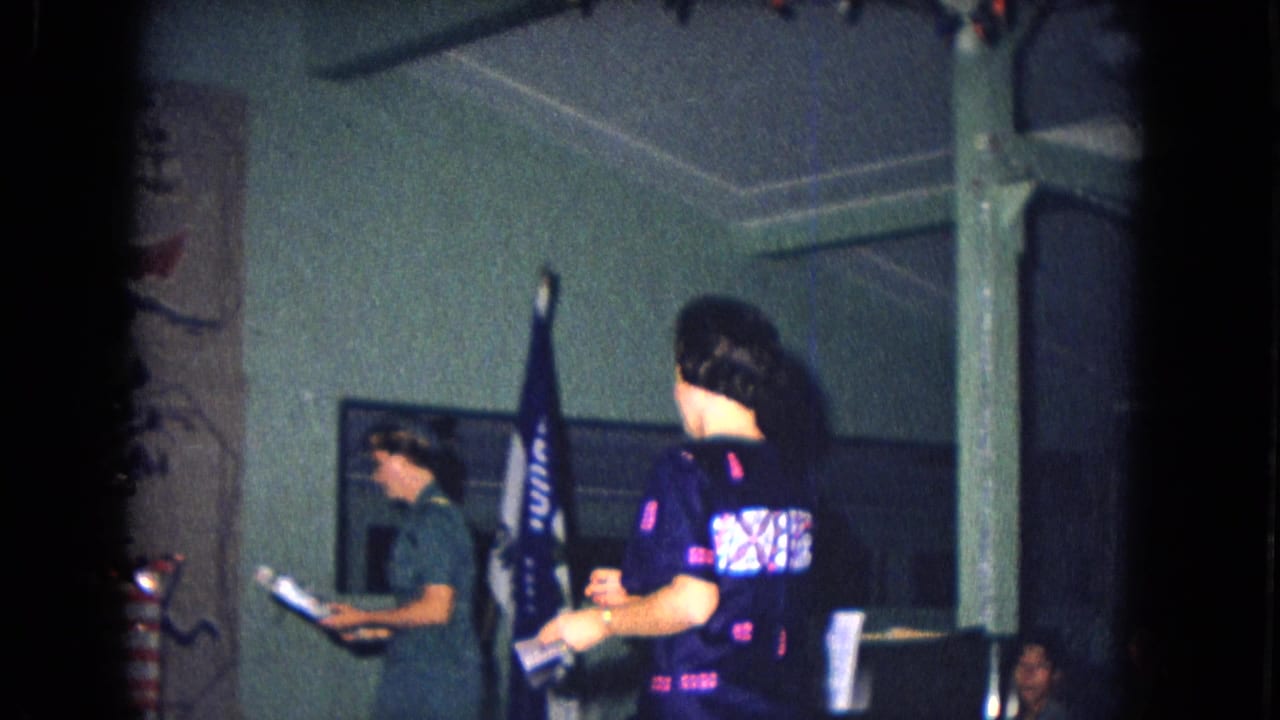 Adults stand inside a decorated room near a flag and wall hanging during a holiday gathering.