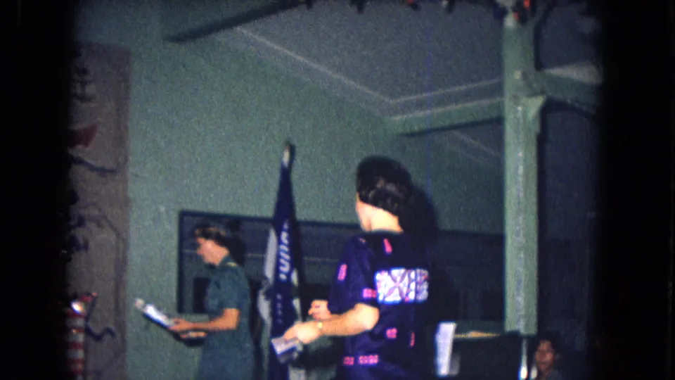 1967 New Mexico 8mm frame showing adults in an interior room with a flag, wall hanging, and holiday decorations nearby.
