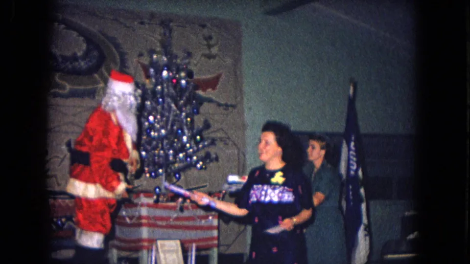 1967 New Mexico home-movie frame of Santa Claus interacting with an adult near a small decorated tree and holiday table.
