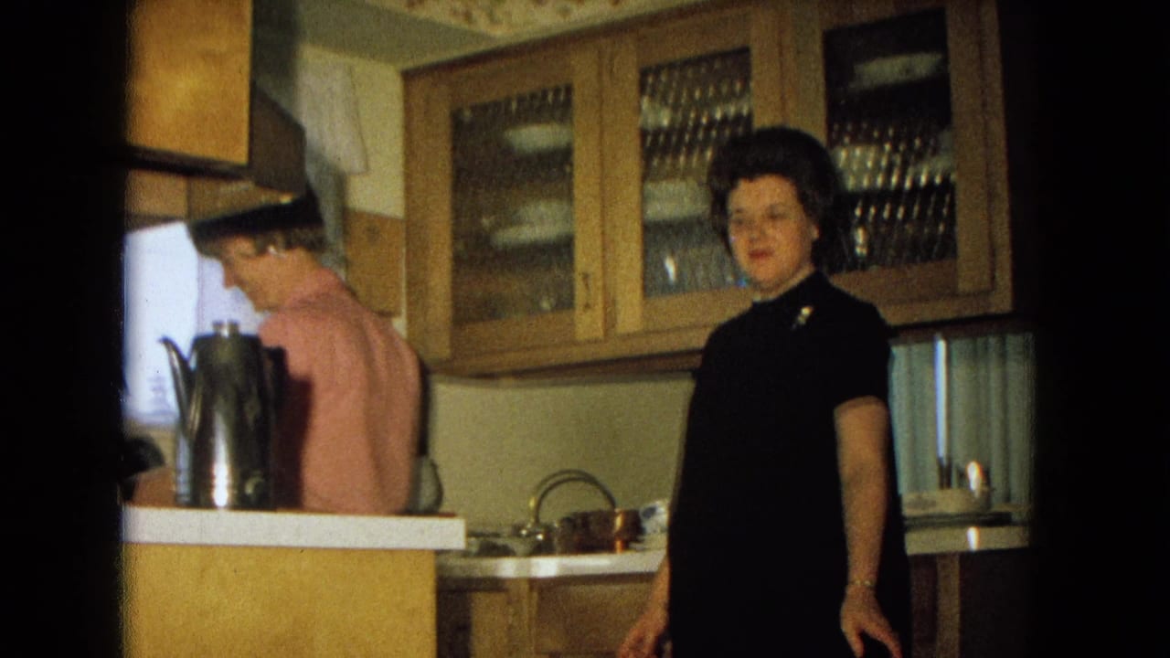 In a 1967 New Mexico kitchen, a woman in a black dress looks toward the camera as another woman stands near the counter.