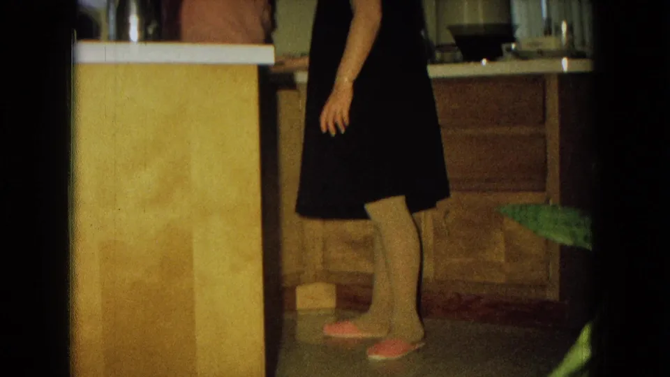 A woman in a dark dress walks through a 1967 New Mexico kitchen with counter space, drawers, and cabinet fronts visible.