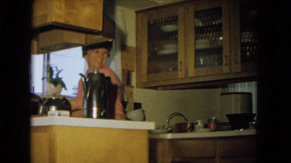A woman in a pink top smiles from behind the counter in a 1967 New Mexico kitchen with glass cabinets and cookware visible.