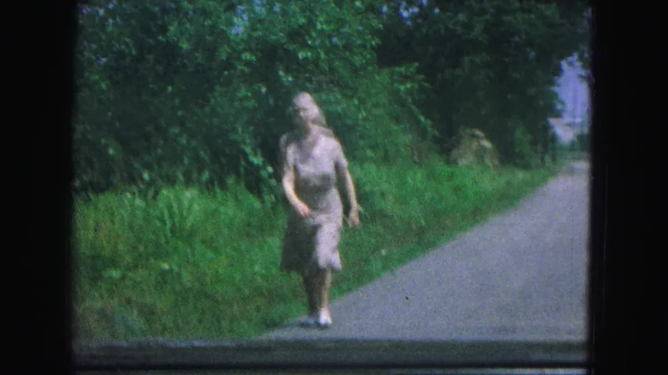 A closer 8mm frame shows the woman in a light dress and white shoes walking beside the paved road in New Mexico.