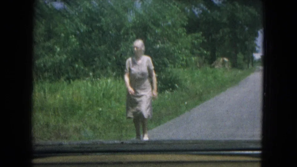 In 1967 New Mexico home-movie footage, an elderly woman walks toward the camera beside grass and trees, seen through a car window.