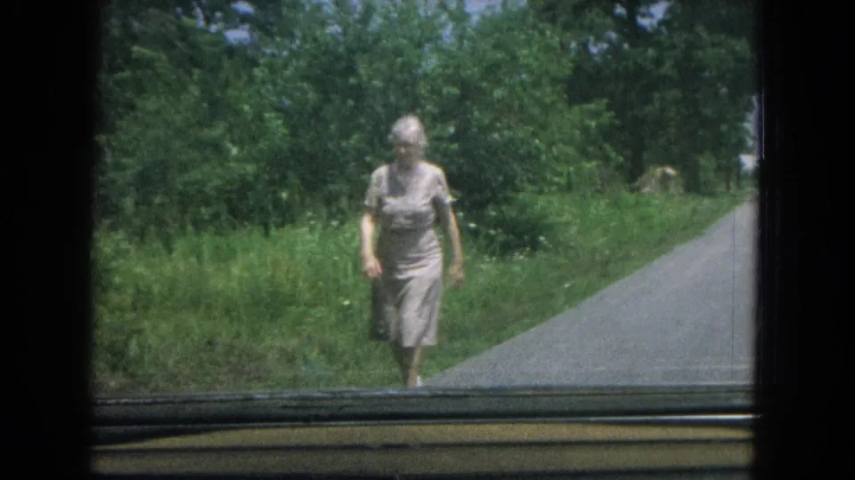 The elderly woman moves closer on the rural driveway, with bright grass and dense trees visible through the car-window frame.