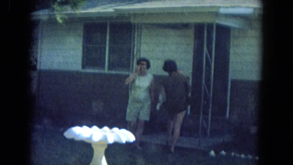 In 1967 New Mexico home-movie footage, two women are near a suburban porch while a white scalloped birdbath sits in the yard.