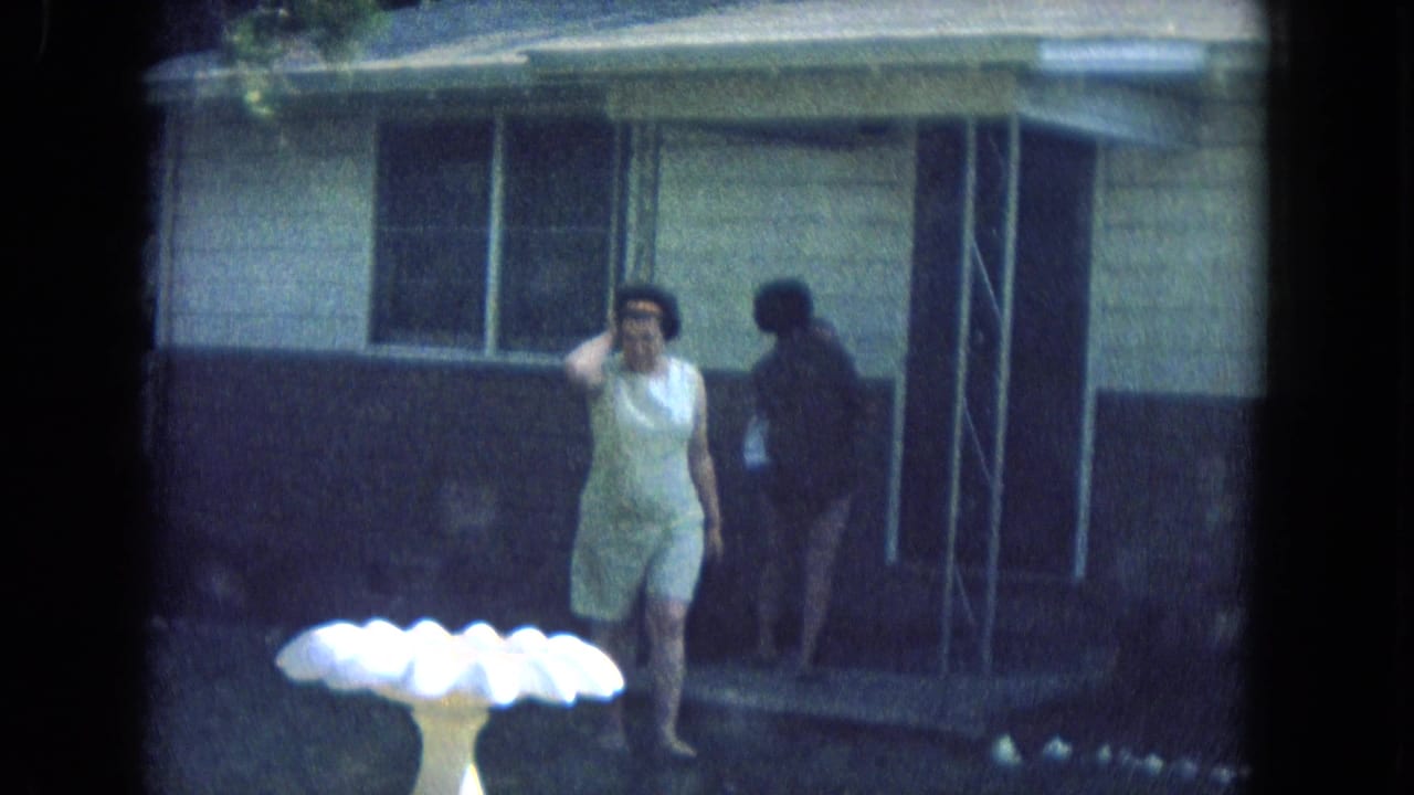 In the New Mexico yard, a woman in a light sleeveless dress adjusts her hair as another woman stands in shadow near the porch.