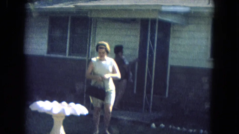 A woman in a light outfit and headband walks toward the camera from the house, holding a dark bag, while another figure is near the porch.
