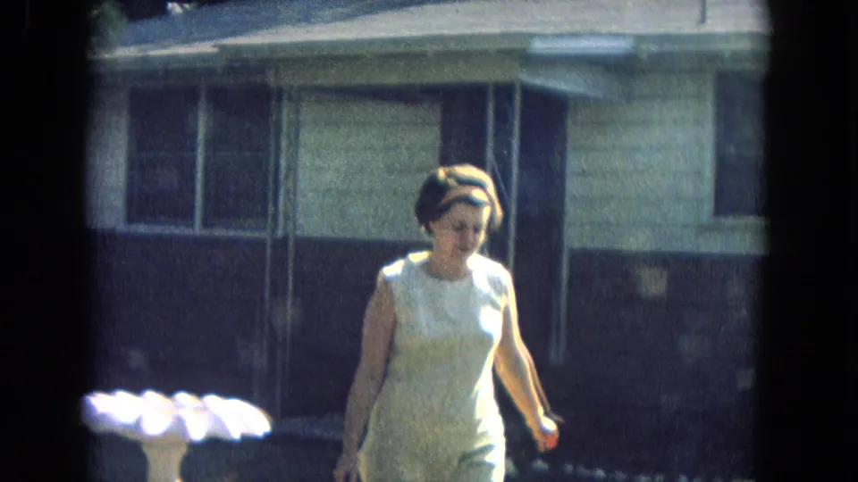 The woman in a light sleeveless dress moves close to the camera, with the porch, windows, and white birdbath still visible behind her.