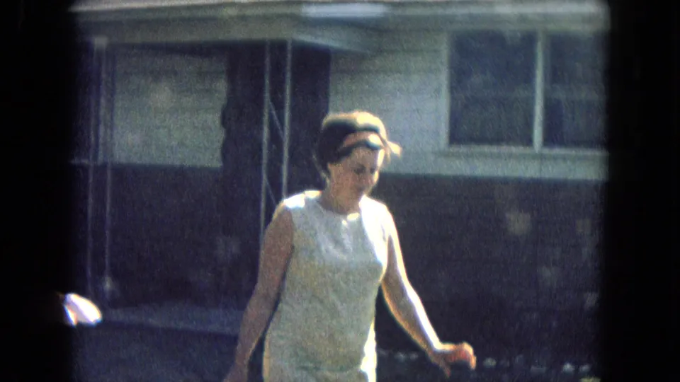 1967 8mm frame of a woman in a light sleeveless dress and headband walking in bright sun near a residential exterior.