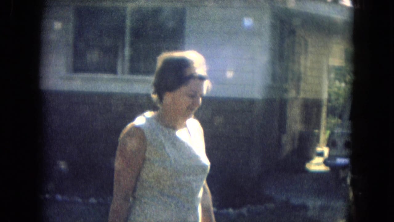 Close 1967 home-movie view of a woman in a light sleeveless dress walking beside a residential facade in strong daylight.
