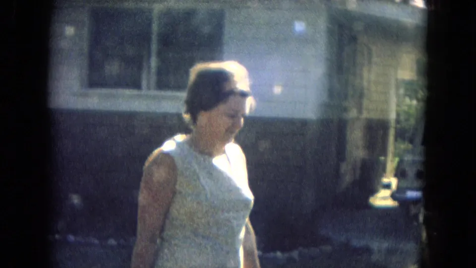 Close 1967 home-movie view of a woman in a light sleeveless dress walking beside a residential facade in strong daylight.