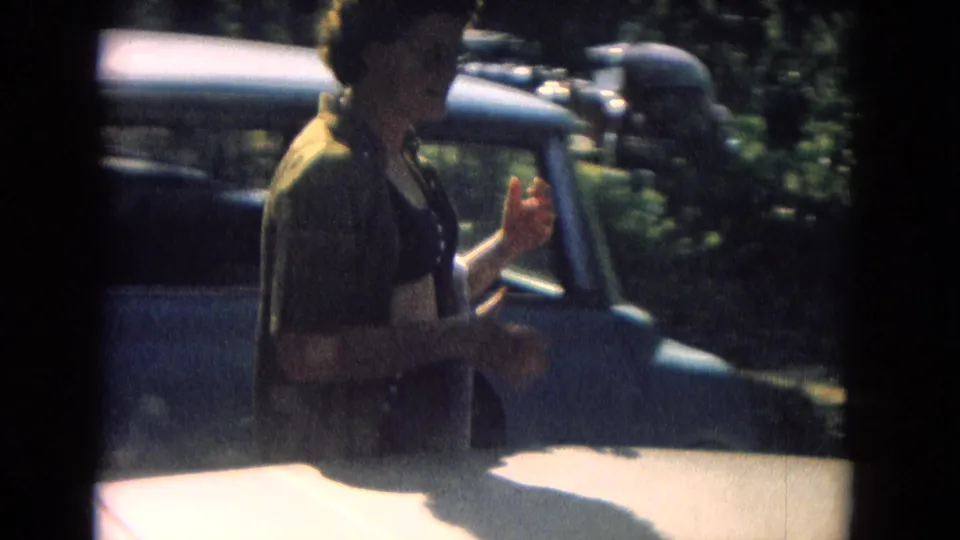 1967 8mm frame showing a woman in a dark top passing an older car, with trees and shadowed foreground visible.