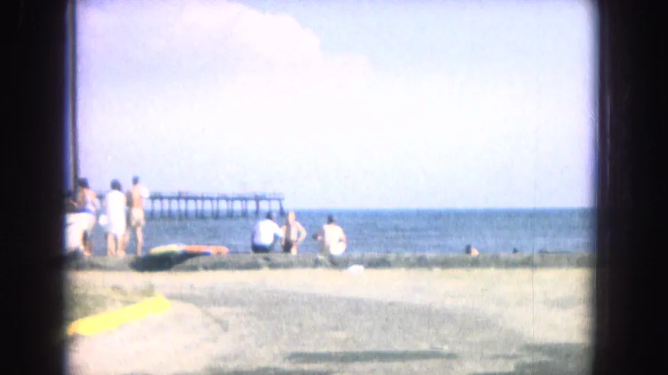 1967 home-movie beach view with people standing and sitting near the shoreline, open water, and a distant pier under a bright sky.
