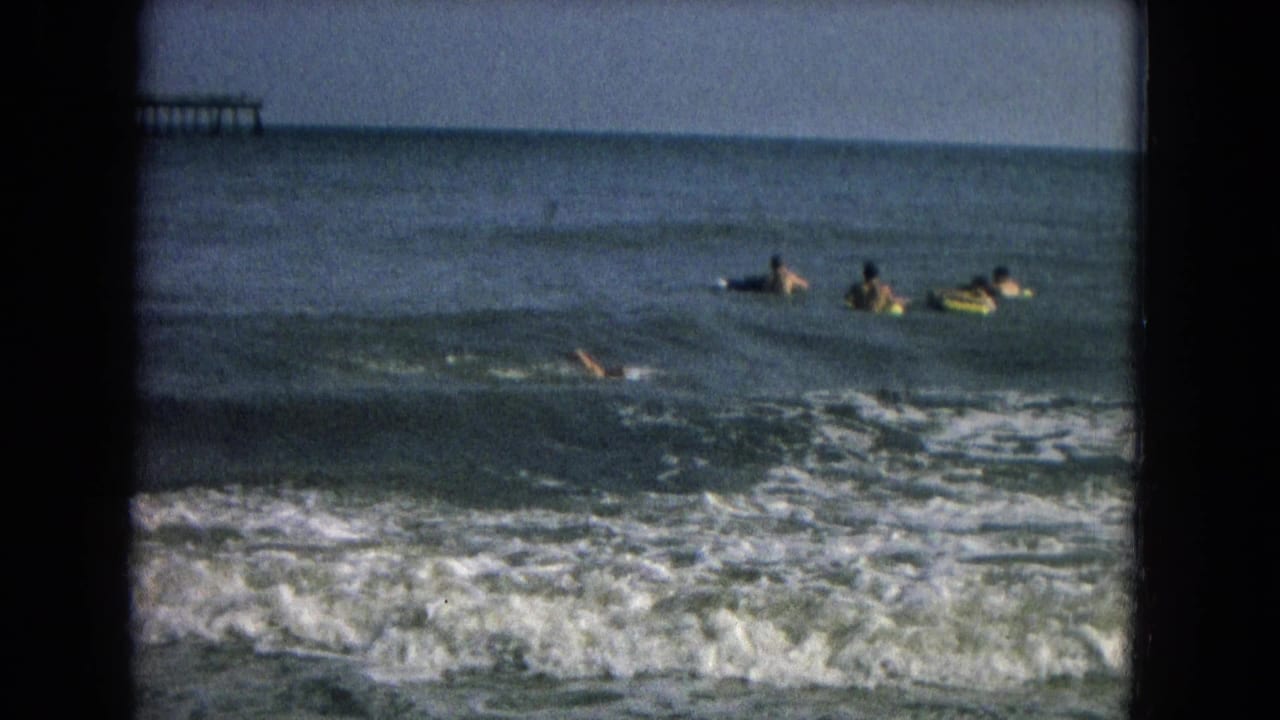 Several swimmers float beyond breaking surf with a pier-like structure in the distance.