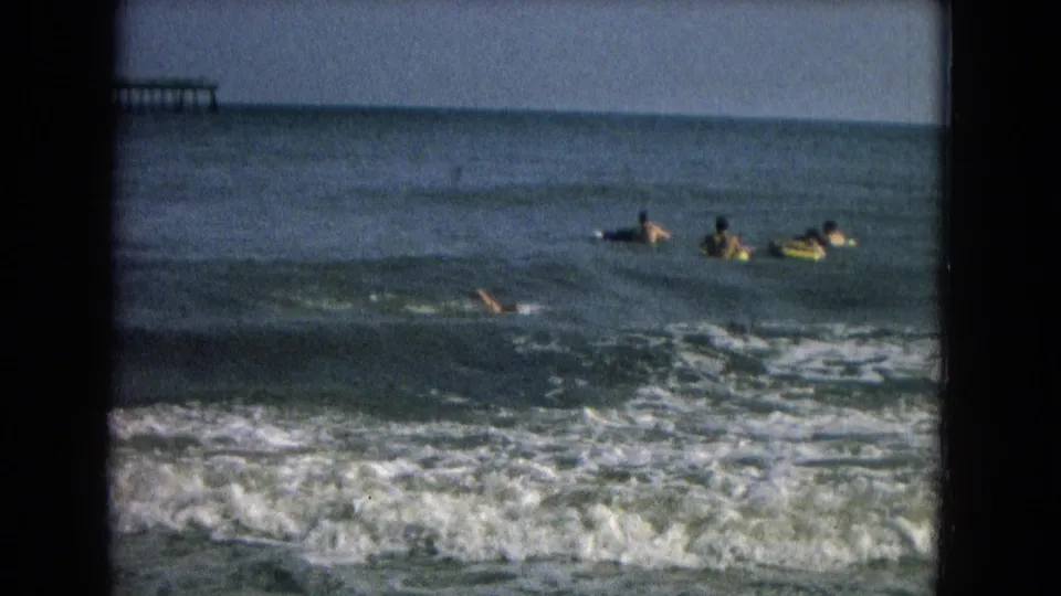 1967 8mm frame showing swimmers spread across open water, white surf in the foreground, and a distant pier-like structure on the horizon.