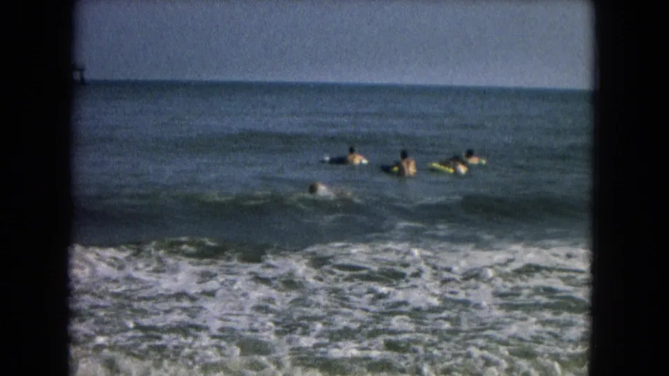 1967 8mm frame of several people floating and paddling beyond the shore break, with foamy surf across the lower frame.